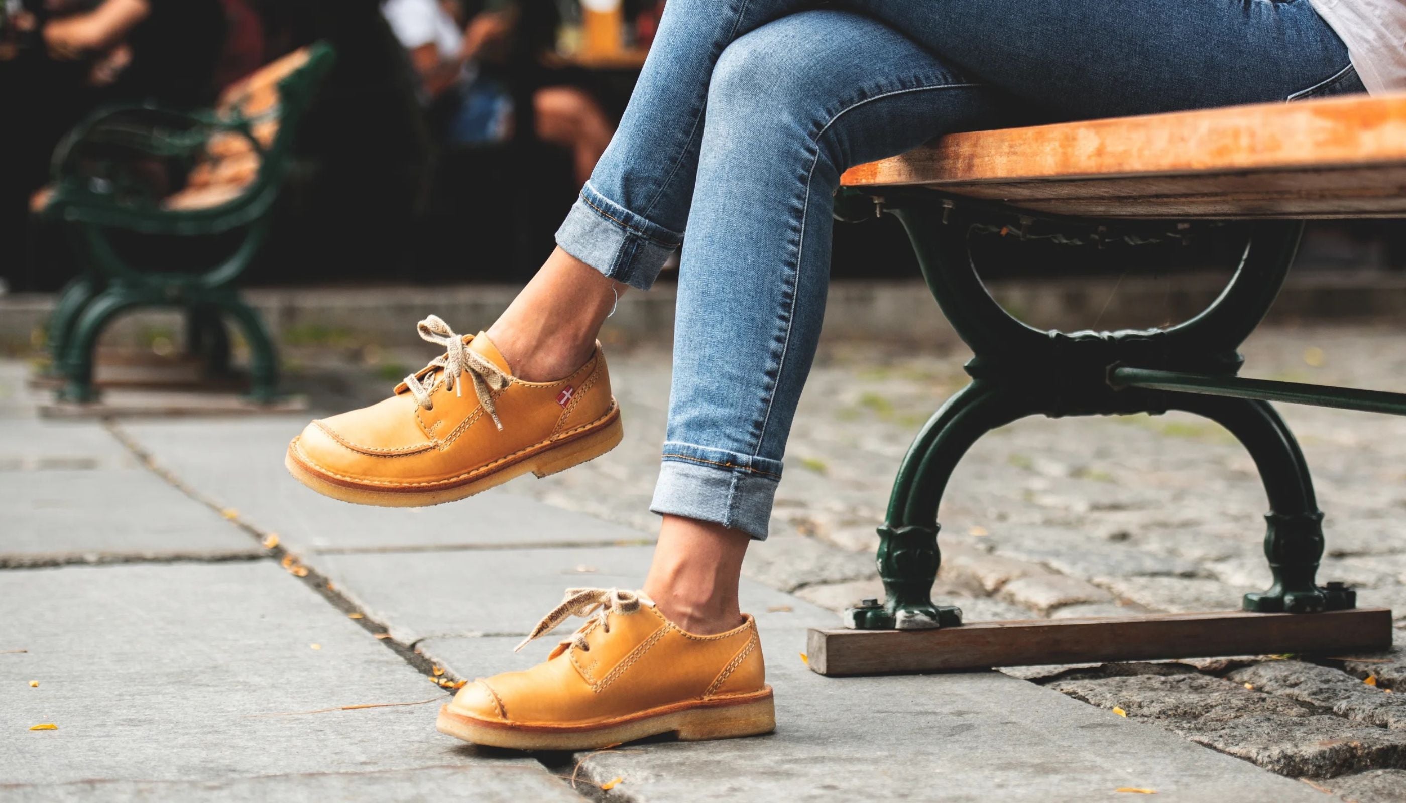 A person sitting on a wooden park bench wearing Duckfeet Fyn moc-toe lace-up shoes and blue jeans, photographed outdoors on a stone pavement with a blurred background.